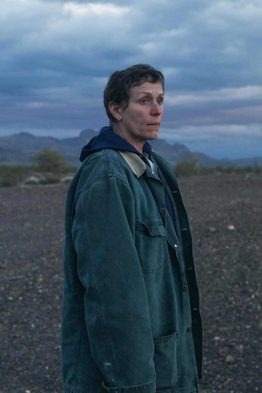 Frances McDormand stands in front of a barren landscape in a work coat