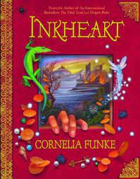Cover of book "Inkheart." Book si red with gol dletters and decorative binding, and features an image of seaside cliffs with a castle. Around the image is a scattering of leaves, gems and pebbles, with a bright green lizard. A hand is crawling out from the image onto the red cover of the book.