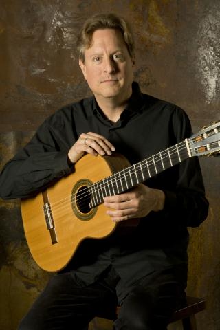 White man with short hair sits on a stool holding an acoustic guitar.