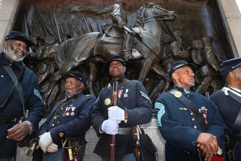 Five black soldiers in uniform and carrying rifles stand at the foot of a war monument