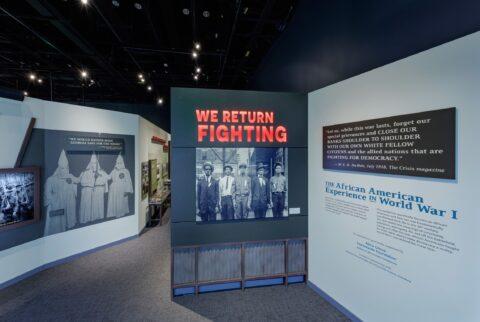 Photo of a museum exhibit entitled We Return Fighting. Black men are standing in the photo.  In the rear of that is a photo of the Ku Klux Klan.