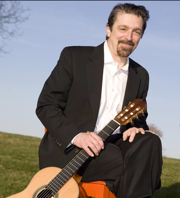 white man with beard sitting on chair outside near green hill holding a guitar