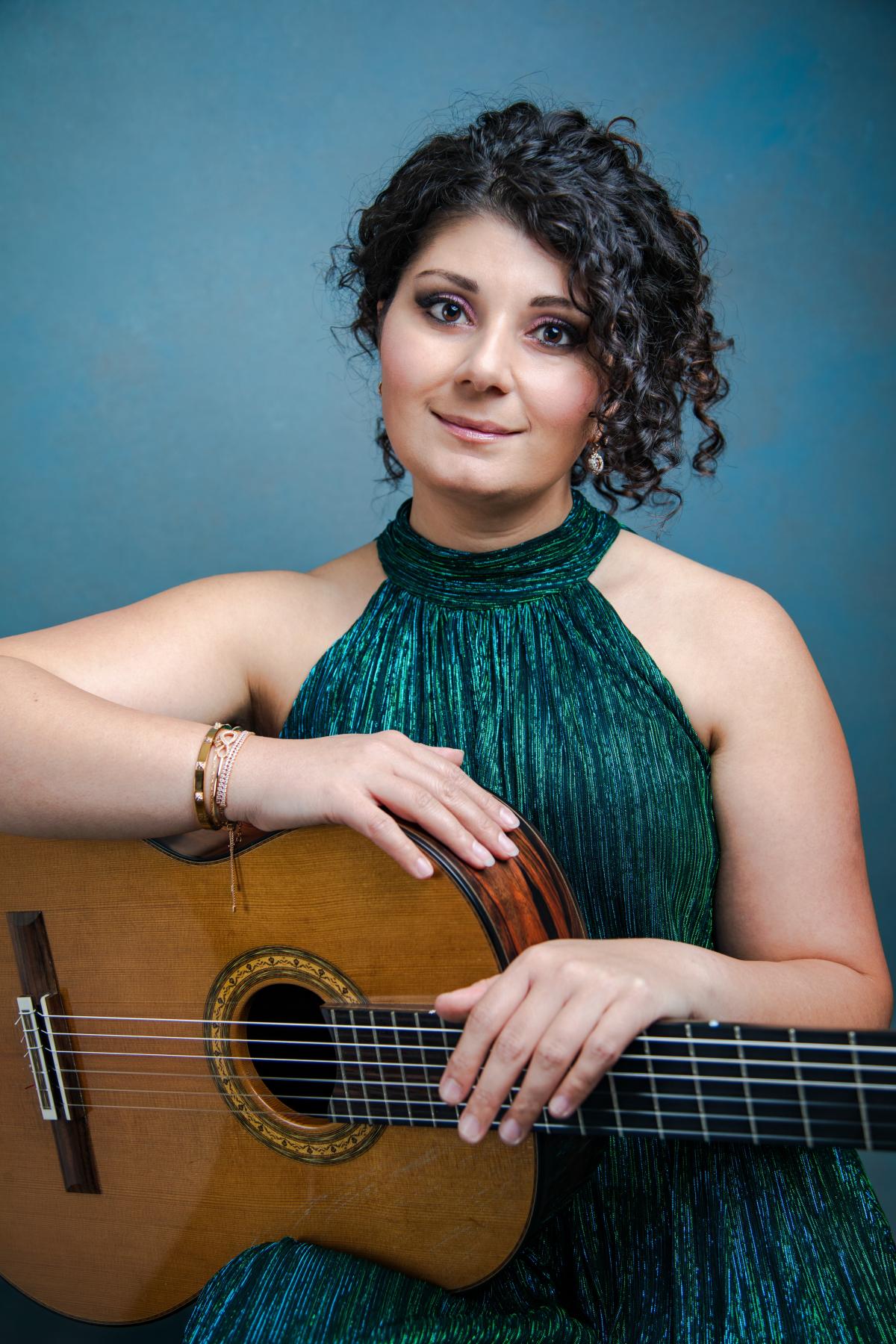 White woman with guitar wearing green formal dress sitting against a teal blue wall.