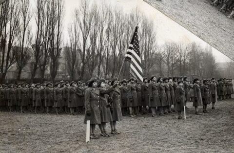 A troop of black women soldiers in uniform hold an American flag in a field